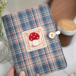 Könnte beinhalten: Ein blau kariertes Notizbuch mit einem roten Pilz-Applikation. Das Notizbuch hat einen Knopfverschluss und einen dekorativen Aufnäher mit dem Text "Hobbyhorse". Der Einband ist aus Stoff.