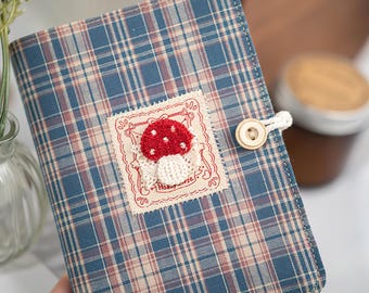 May include: A blue plaid journal with a red mushroom applique. The journal has a button closure and a decorative patch with the text "Hobbyhorse". The cover is made of fabric.