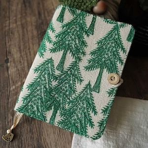 Puede incluir: Una cubierta de diario con un patrón de pinos bordados en verde sobre un fondo beige. La cubierta se cierra con un botón de madera y una cuerda. Una cinta dorada está unida al diario.