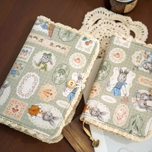 May include: Two fabric-covered journals featuring a light green grid pattern with illustrations of rabbits, teapots, and flowers in scalloped frames. Each journal is edged with cream-colored lace trim and secured with a small wooden button closure.
