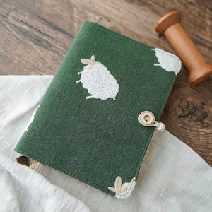 Könnte beinhalten: Ein grünes Stoffjournal mit weißen, gestickten Schafmotiven. Das Journal hat einen Holzknopfverschluss und passende gestickte Schafe an den Ecken. Eine Holzspule ist ebenfalls zu sehen.