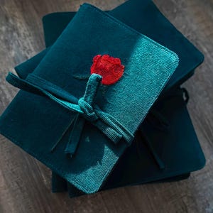 May include: A stack of teal corduroy notebooks tied with a teal ribbon. Each notebook features a red rose embroidery on the front. The notebooks are stacked on top of each other, with the top one in focus.