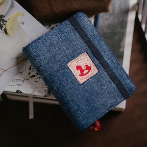 May include: A rectangular notebook with a dark blue denim fabric cover and a vertical black elastic band closure. A square patch is centered on the front, featuring a red rocking horse illustration and the text Hobbyhorse in red ink.