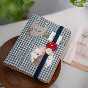 May include: A fabric-covered notebook featuring a blue and white gingham pattern. The cover includes a dark blue elastic band, a silver metal paperclip, and a small embroidered red and white mushroom appliqué attached to the elastic strap.