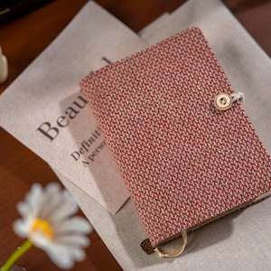 Op de afbeelding: Een rood en crèmekleurig geweven notitieboek met een knoopsluiting. Het notitieboek ligt op een linnen doek met het woord "Beautiful" erop gedrukt. Een madeliefje staat in de linker benedenhoek.