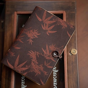 May include: A dark brown fabric journal cover featuring a repeating pattern of reddish-brown bamboo leaves. The cover is secured with a small, dark brown button and a matching fabric loop closure, providing a simple and functional design.