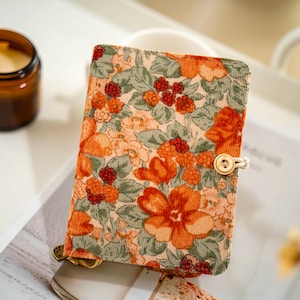 Puede incluir: Un diario de pana con un estampado floral en naranja, rojo y verde. Tiene un cierre de botón de madera con una cuerda crema. La cubierta presenta un patrón repetido de flores y bayas.