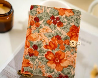 May include: A corduroy journal with a floral pattern in orange, red, and green. The journal has a wooden button closure with a cream-colored string. The cover features a repeating pattern of flowers and berries.