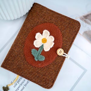 May include: A brown fabric journal cover with a woven daisy applique. The daisy has white petals and a yellow centre, with green leaves. The cover has a button closure and a decorative charm.