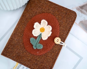 May include: A brown fabric journal cover with a woven daisy applique. The daisy has white petals and a yellow center, with green leaves. The cover has a button closure and a decorative charm.