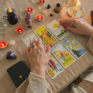 May include: A tarot card reading setup with lit candles, crystals, and a deck of cards. The cards are laid out on a beige surface, with hands hovering over them. The cards feature colorful illustrations and symbols, suggesting a focus on divination and spirituality.