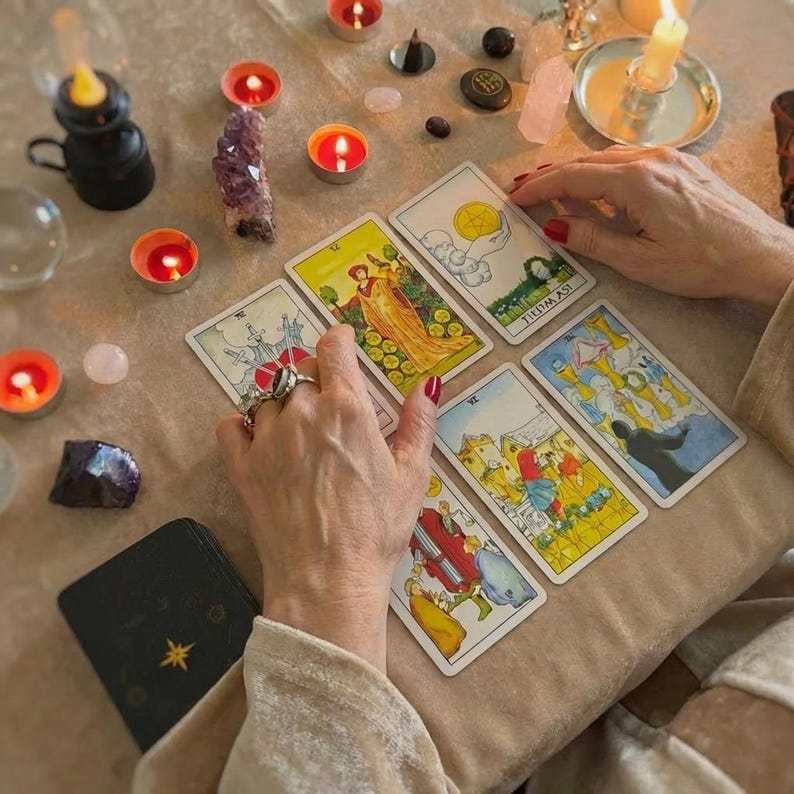 May include: A tarot card reading setup with lit candles, crystals, and a deck of cards. The cards are laid out on a beige surface, with hands hovering over them. The cards feature colorful illustrations and symbols, suggesting a focus on divination and spirituality.