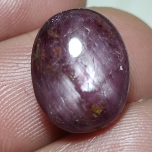 May include: A polished, oval-shaped gemstone with a deep purplish-red hue. The surface has a glossy sheen with subtle variations in color and small inclusions. The gem is held between fingers, showcasing its size and shape.