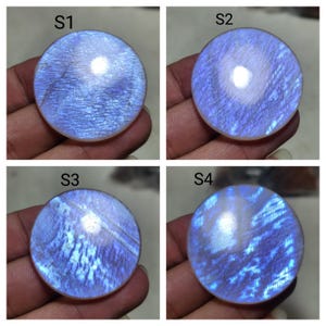 May include: Four circular moonstone cabochons labeled S1, S2, S3, and S4. Each polished gemstone displays a translucent, pale blue body with a distinct adularescence effect, showing internal light reflections and fine, linear surface textures.