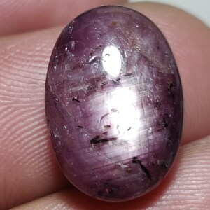 May include: A polished, oval gemstone in a deep purplish-red colour. The stone has a smooth, reflective surface with internal inclusions and dark flecks. The gemstone is held in a hand, highlighting its colour and texture. A close-up view shows the stone's natural beauty.