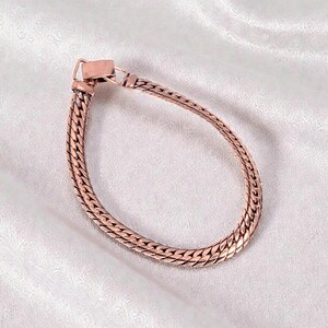 May include: A copper-colored bracelet with a woven chain design. The bracelet features a rectangular clasp and is displayed on a white, textured surface. The jewelry piece is a stylish accessory.