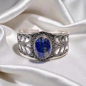May include: Silver cuff bracelet featuring an oval, deep blue gemstone. The gemstone is set in an ornate, filigree-style frame. The bracelet has a wide band with decorative openwork patterns. The background is a white, silky fabric.