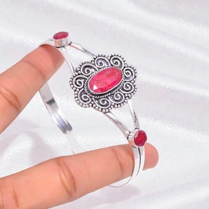 May include: A silver cuff bracelet with a central oval ruby gemstone, framed by an elaborate, swirling pattern. Two smaller ruby stones are set on the bracelet's ends. The bracelet is displayed against a white backdrop.