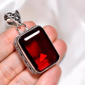 May include: A rectangular pendant featuring a large, deep red gemstone set in a decorative silver frame with a filigree bail. The stone has a faceted surface that reflects light, and the metal setting displays intricate, textured detailing.