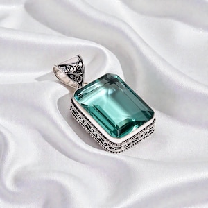 May include: A silver pendant featuring a large, rectangular, light-blue gemstone. The pendant has an ornate, filigree design around the gemstone and on the bail. The gemstone is a vibrant, cool colour, and the silver setting adds contrast.