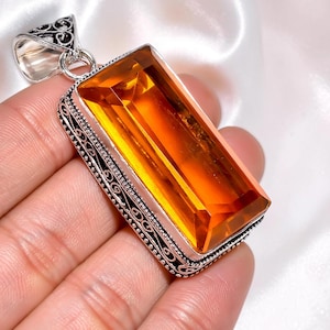 May include: A rectangular pendant featuring a large, faceted orange citrine gemstone set in a silver frame with intricate scrollwork and beaded detailing along the edges, complete with a decorative silver bail at the top for hanging.