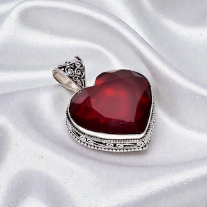 May include: A heart-shaped pendant with a large, faceted, deep red gemstone. The pendant is set in a silver-colored metal with ornate detailing around the edges and bail. The gemstone is the focal point, catching the light.