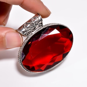 May include: A large, oval-shaped pendant with a vibrant red gemstone. The gemstone is set in a silver-colored metal frame with intricate detailing. The pendant is held by a finger.