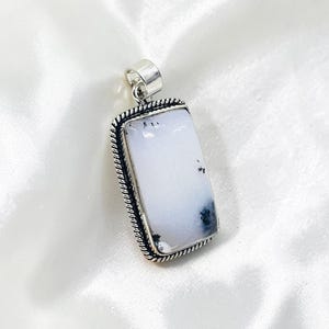 May include: A rectangular pendant with a white stone, featuring black and grey inclusions. The stone is set in a silver-coloured metal frame with a twisted rope design around the edges. A silver loop is at the top for a chain.