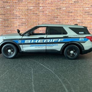 May include: A silver and blue Jefferson County Sheriff SUV model. The vehicle has black wheels and a blue stripe with the word "SHERIFF" in white. The text "TO PROTECT AND SERVE" is also visible.