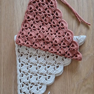 May include: A handmade, two-tone crochet bonnet in a soft, muted color palette. The top section is a dusty rose, while the bottom is a creamy white. The bonnet features a scalloped edge and ties for a secure fit. The intricate openwork design adds a delicate touch.