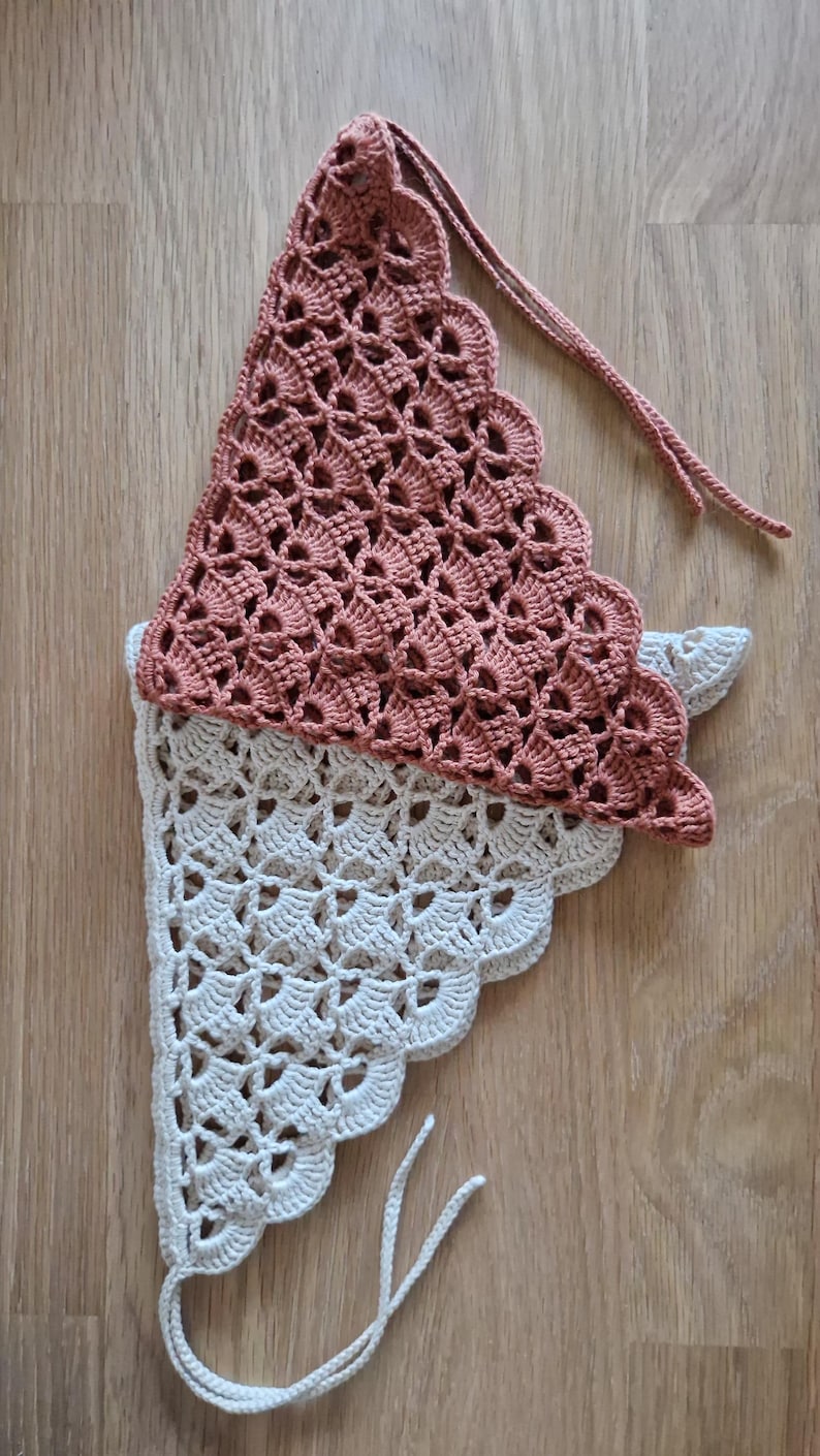 May include: A handmade, two-tone crochet bonnet in a soft, muted color palette. The top section is a dusty rose, while the bottom is a creamy white. The bonnet features a scalloped edge and ties for a secure fit. The intricate openwork design adds a delicate touch.