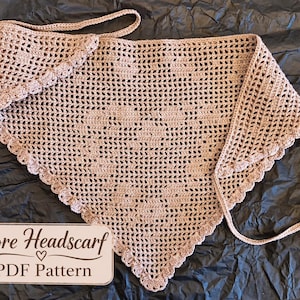 Puede incluir: Un pañuelo triangular de ganchillo en color beige claro con un patrón de malla calada, un motivo de corazón en el centro y un borde festoneado. Una etiqueta rectangular en la esquina muestra el texto Cuore Headscarf PDF Pattern.