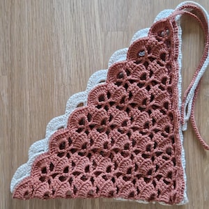 May include: A handmade crochet bandana in a triangular shape. The bandana features a scalloped edge and is made of two colors: a light beige and a muted reddish-brown. The crochet work has an open, lacy design, and it has two long ties.