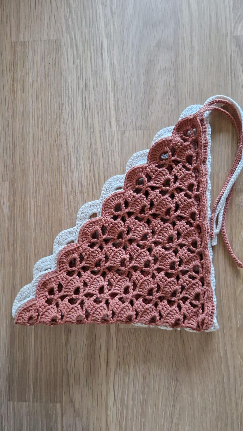 May include: A handmade crochet bandana in a triangular shape. The bandana features a scalloped edge and is made of two colors: a light beige and a muted reddish-brown. The crochet work has an open, lacy design, and it has two long ties.