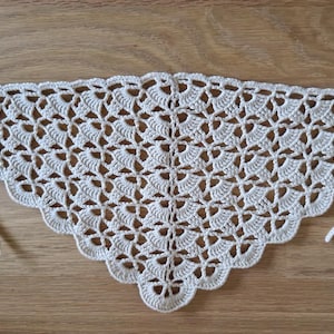 May include: Cream-colored, crocheted triangle shawl with a scalloped edge. The openwork design features a repeating floral pattern. The shawl has two long ties at the corners for securing around the neck or shoulders. The shawl is displayed on a wooden surface.