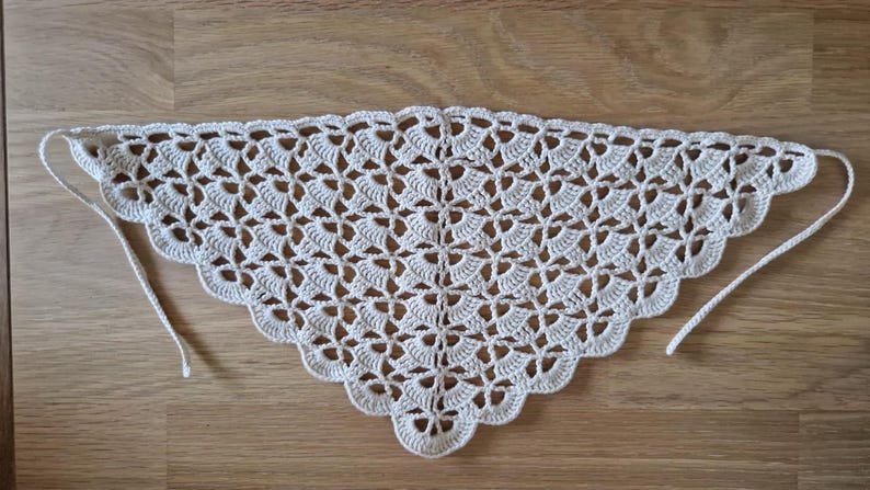 May include: Cream-colored, crocheted triangle shawl with a scalloped edge. The openwork design features a repeating floral pattern. The shawl has two long ties at the corners for securing around the neck or shoulders. The shawl is displayed on a wooden surface.