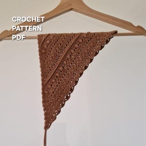 Può includere: Una sciarpa triangolare all'uncinetto marrone appesa a una gruccia di legno. La sciarpa presenta un motivo traforato dettagliato e un bordo smerlato. Il testo "CROCHET PATTERN PDF" è visualizzato in bianco.