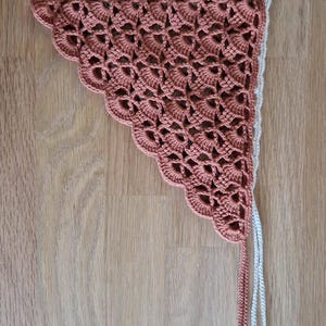 May include: Two handmade crochet triangle scarves. The top scarf is off-white, and the bottom scarf is a muted coral color. Both scarves have a scalloped edge and long, braided ties.