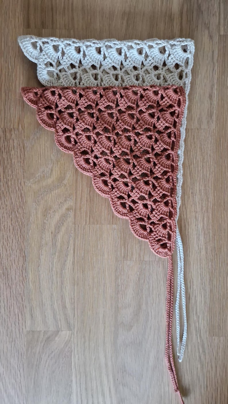 May include: Two handmade crochet triangle scarves. The top scarf is off-white, and the bottom scarf is a muted coral color. Both scarves have a scalloped edge and long, braided ties.