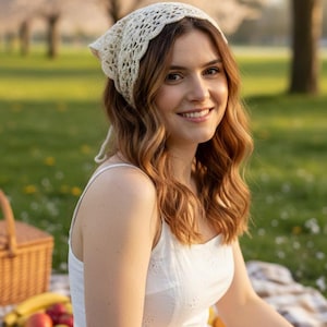 May include: A woman wearing a white eyelet dress and a cream-colored crocheted headscarf. She is sitting on a picnic blanket with a basket of fruit nearby. The background features a grassy area and trees.