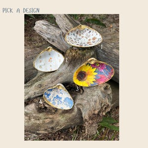 May include: Four decorative seashell dishes with gold trim, each featuring a unique design. Designs include floral patterns, a sunflower, and a blue and white toile print. The text "Pick a Design" is at the top left.