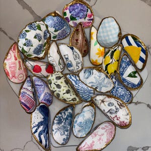 May include: A collection of decorative seashells, each hand-painted with unique designs. The shells feature floral patterns, nautical themes, and other artistic motifs, with gold-painted edges. The designs include hydrangeas, lemons, and strawberries.