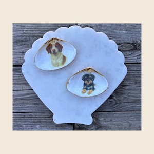 May include: Two seashell trinket dishes with dog portraits, set on a white, scalloped marble tray. One shell features a brown and white dog, the other a black and brown dog. The shells have gold-painted edges.