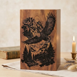 May include: A rectangular wooden urn featuring a dark, laser-engraved illustration of a bald eagle in flight above a dense pine forest and jagged mountain peaks. The natural wood grain is visible across the surface of the upright container.