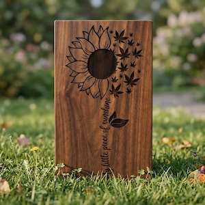 Könnte beinhalten: Rechteckige Holzplakette mit einem Sonnenblumen- und Cannabisblatt-Design. Die Sonnenblume befindet sich links und geht in Cannabisblätter auf der rechten Seite über. Der Satz "my little piece of sunshine" ist vertikal eingraviert. Das Holz hat eine natürliche braune Farbe.