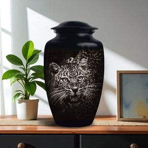 May include: Black cremation urn with a detailed grayscale image of a snarling leopard. The urn has a rounded shape with a lid. The leopard's face is the focal point, showcasing its teeth and intense gaze. A decorative item for memorial purposes.