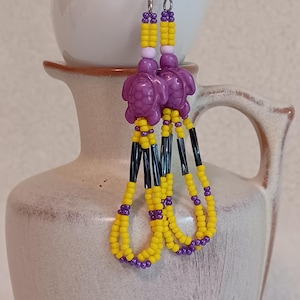 May include: Handmade beaded earrings featuring purple turtle charms. The earrings have yellow and purple beads, with black accents. The earrings are hanging in front of a white ceramic vase.