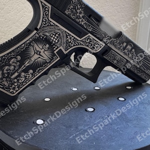 Op de afbeelding: Een zwart semi-automatisch pistool met gedetailleerde lasergravures van religieuze afbeeldingen. Het greepgedeelte toont een afbeelding van de kruisiging, terwijl de slede is versierd met bloemmotieven en andere religieuze scènes.