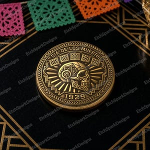 May include: A circular gold-toned metal coin featuring an embossed skull design in the centre. The text "DÍA DE LOS MUERTOS" is engraved along the top edge, and the number "1929" is engraved at the bottom, surrounded by geometric patterns.