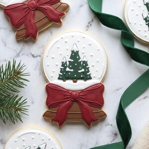 Op de afbeelding: Kerstmis sneeuwbolkoekjes. Elke koekje heeft een wit glazuur met groene bomen en vallende sneeuw. Een rode strik en bruine basis maken het ontwerp compleet. Een groen lint en een dennentak staan ook op de foto.